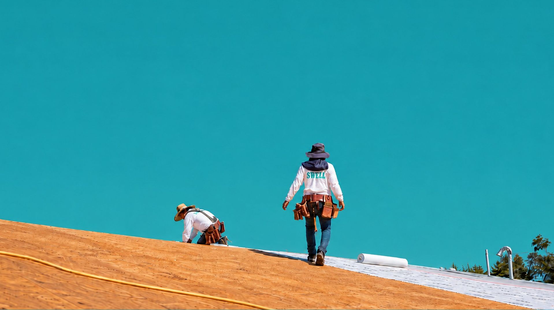 Two Swell roofers laying underlayment on a fresh plywood roof deck under a clear Southern California sky