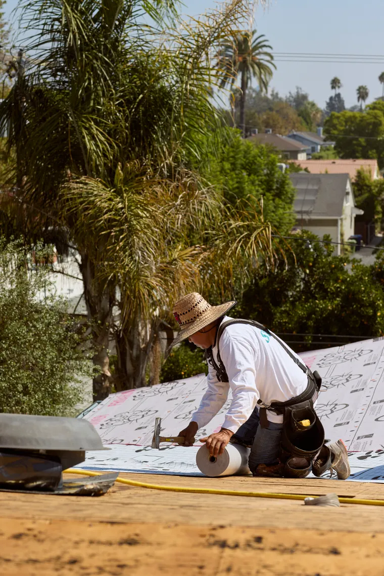 Worker fastening underlayment on a pitched roof with palm trees beyond