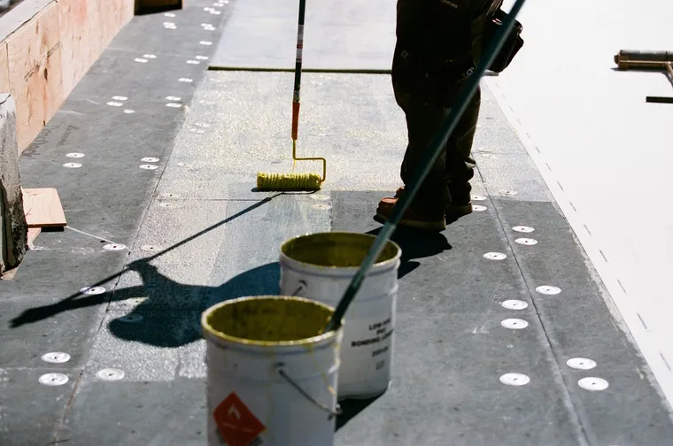 Two rollers spreading bonding adhesive over fastened coverboard