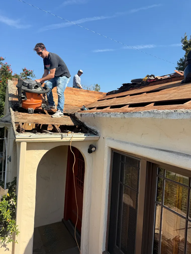 Worker using a shop vacuum on exposed sheathing with clay tiles stacked nearby